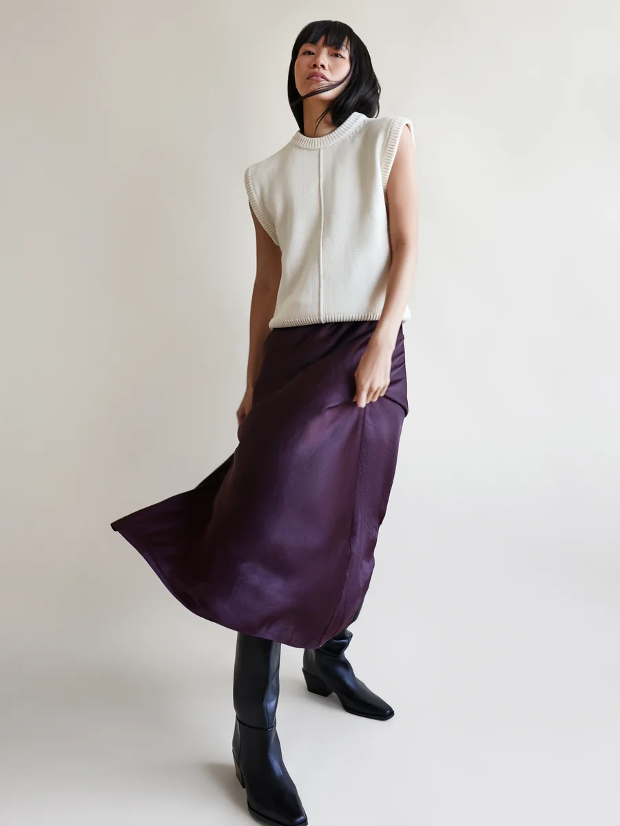 Darin Apinya in a white knit vest and flowing purple skirt against a studio backdrop